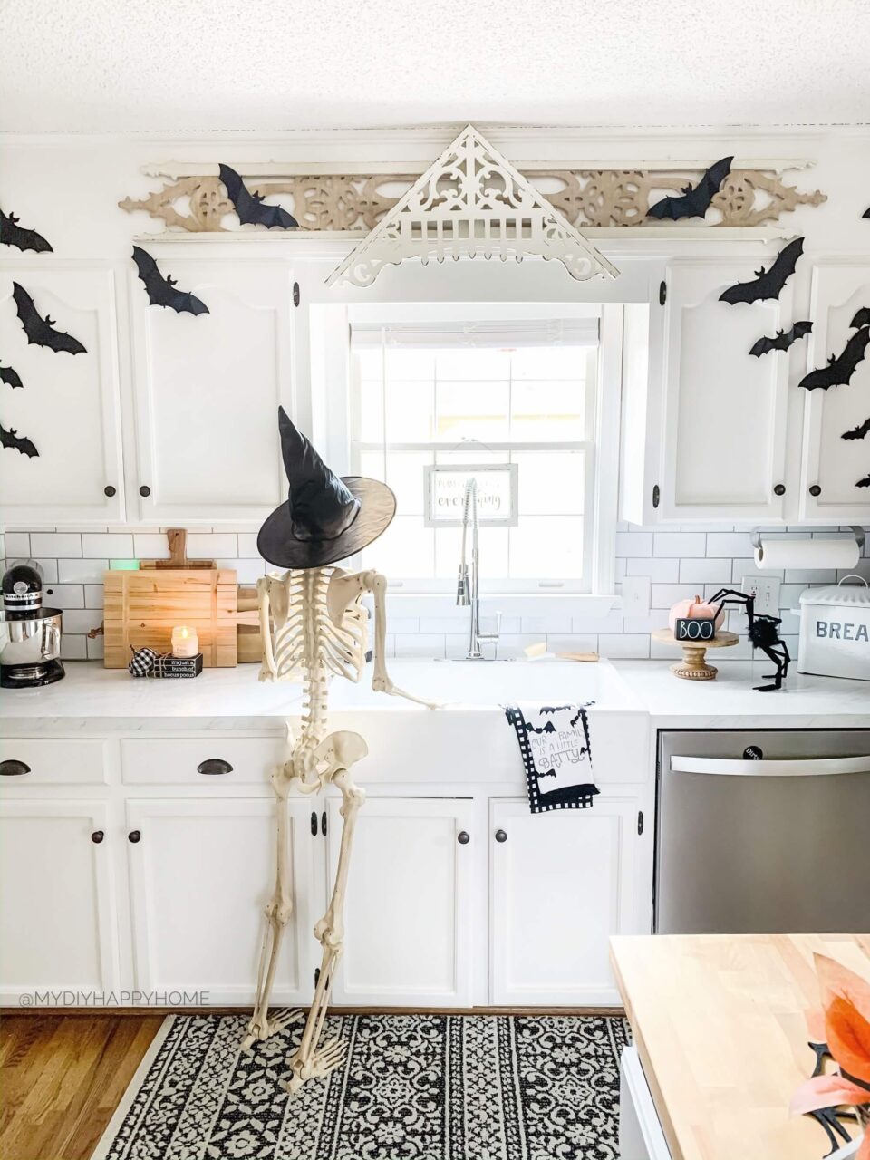 A Halloween-themed kitchen with a skeleton wearing a witch hat standing by the sink, surrounded by bat decals on the cabinets and festive decorations on the counter.