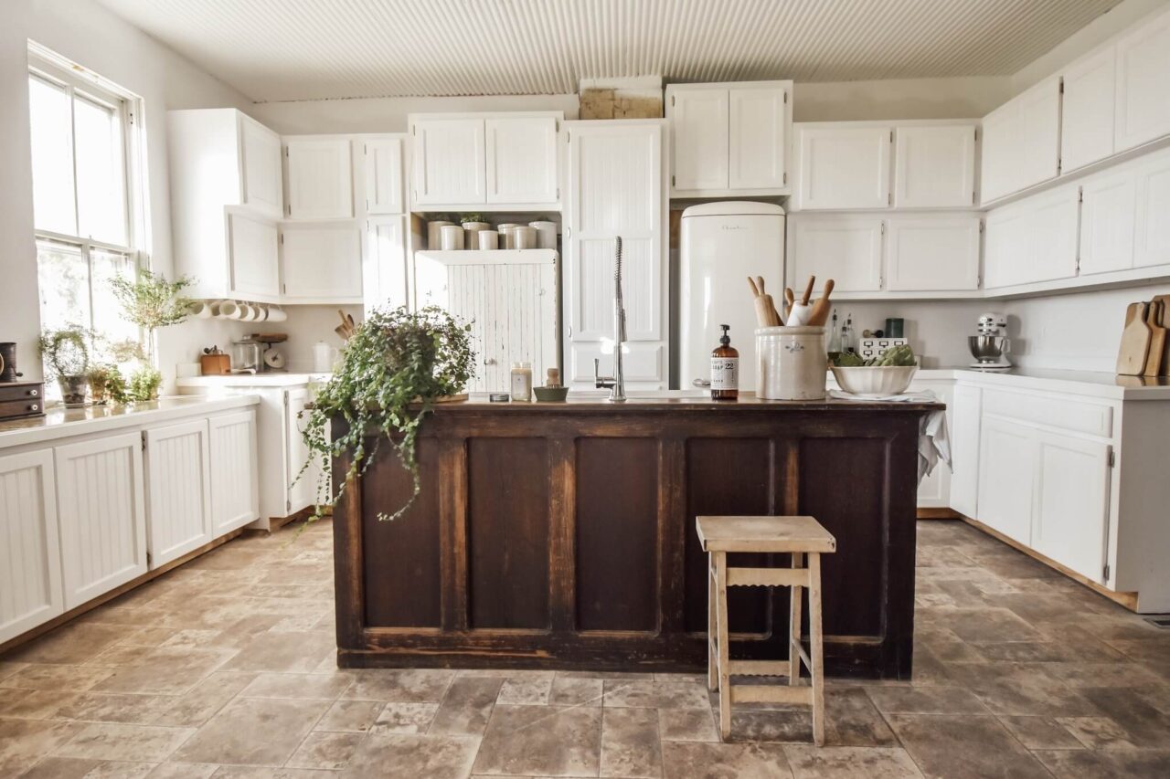 A rustic kitchen with white cabinets, a wooden island, plants, and kitchen tools, featuring a farmhouse sink and vintage refrigerator.