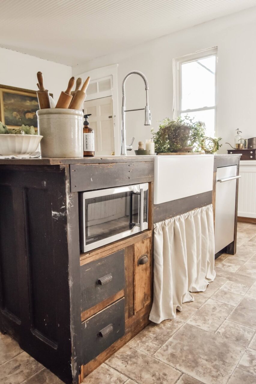 A rustic kitchen island with a white farmhouse sink, microwave, and vintage-style cabinets, featuring rolling pins, a plant, and a dish soap bottle on the countertop.