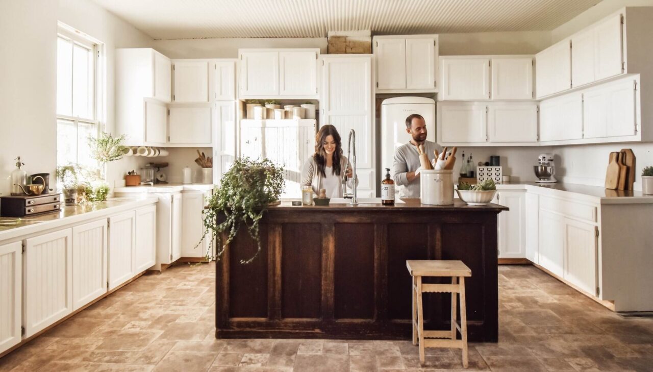 A couple standing at a rustic kitchen island in a bright kitchen with white cabinets, plants, and various kitchen items.
