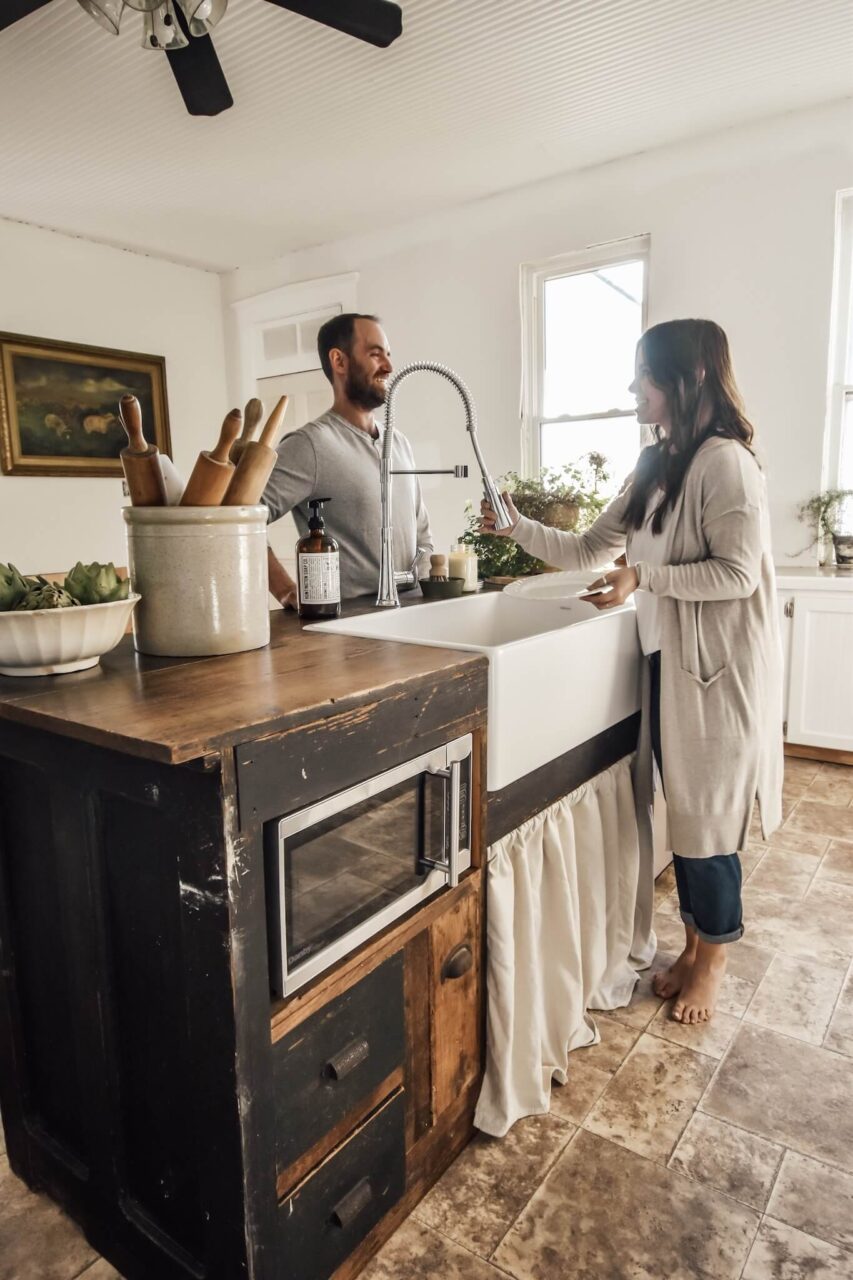 A couple stands at a rustic kitchen island with a farmhouse sink, engaged in conversation and washing dishes.