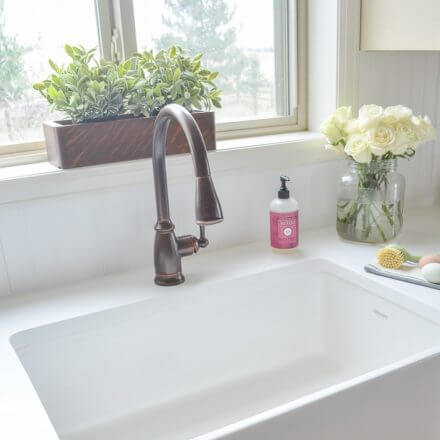 A clean, white kitchen sink with a bronze faucet, a potted plant on the windowsill, and a vase of white roses beside a bottle of soap.