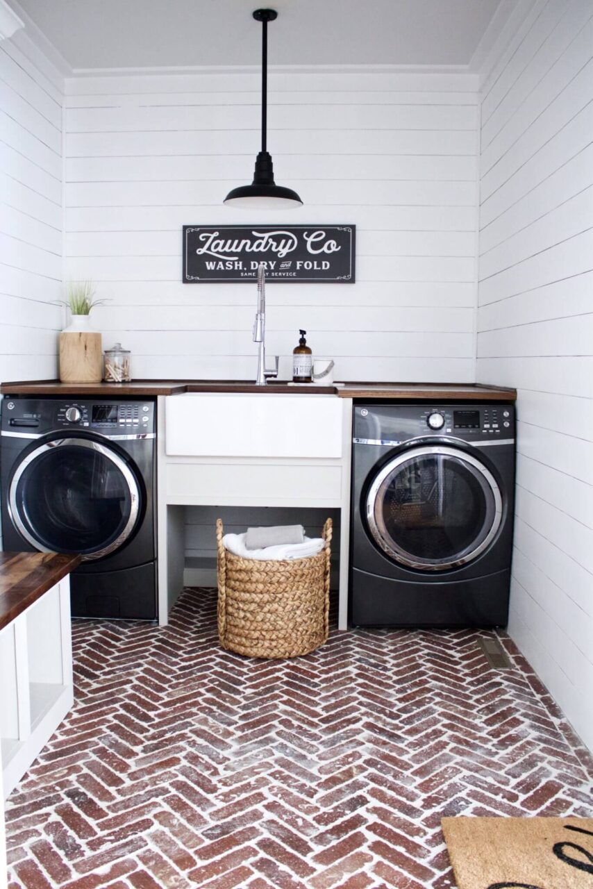 A stylish laundry room with a black washer and dryer, a farmhouse sink, a "Laundry Co" sign, and a woven laundry basket on a herringbone brick floor.