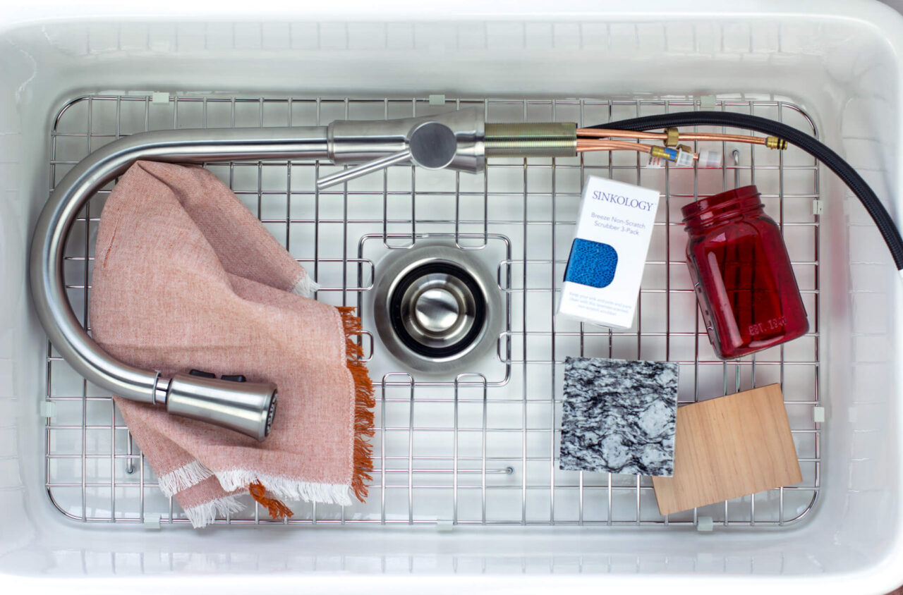 A kitchen sink with a stainless steel faucet, a pink cloth, a box of Sinkology non-scratch scrubbers, a red mason jar, and two material samples.