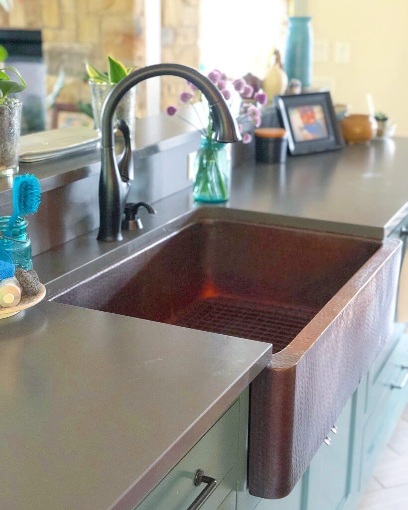 A close-up of a kitchen island with a hammered copper farmhouse sink, black faucet, and various decorative items including a vase with purple flowers and a photo frame.