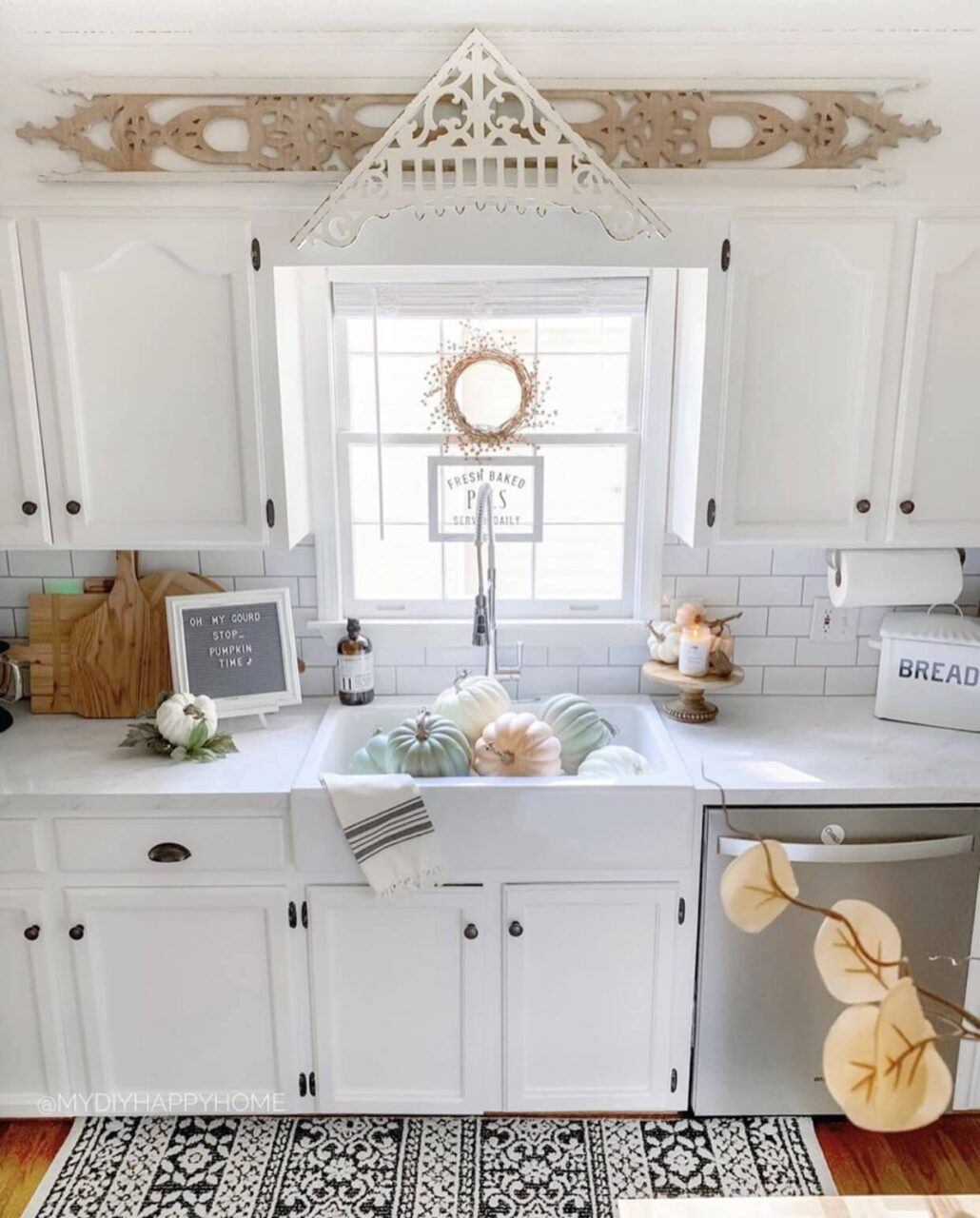 A kitchen decorated for fall with pastel pumpkins in the farmhouse sink, a wreath hanging in the window, and a letter board with a seasonal message on the counter.