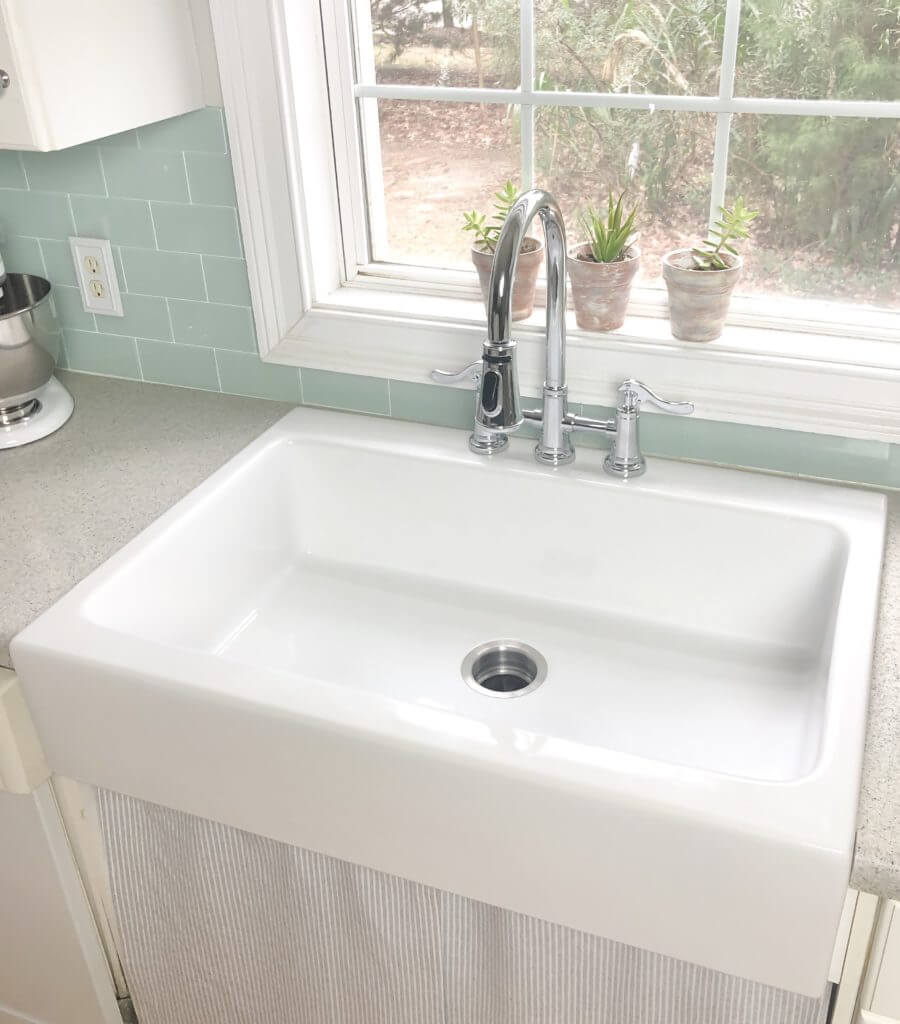 A white farmhouse sink with a chrome faucet, set in a kitchen with a light green tile backsplash and small potted plants on the windowsill.