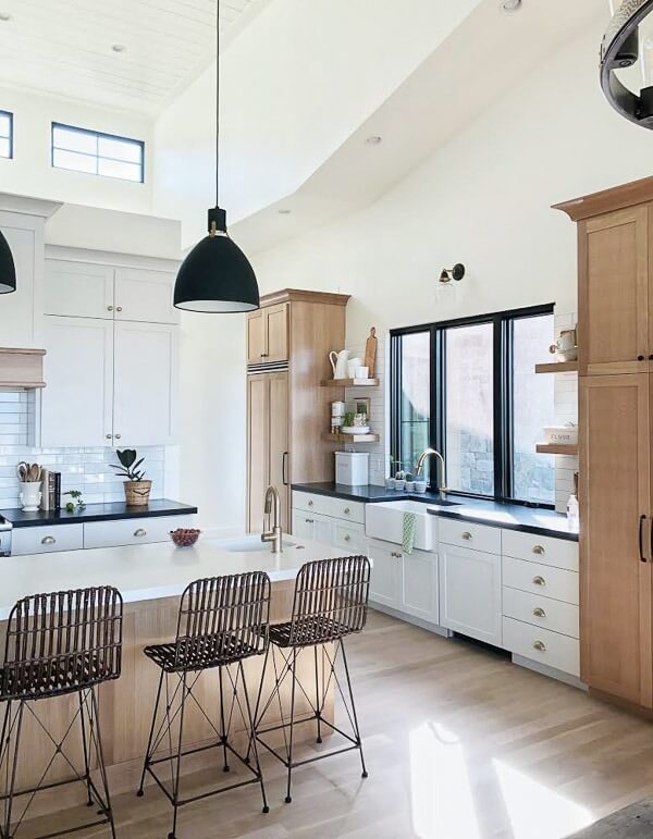 A modern kitchen with white cabinets, a large island with wicker bar stools, pendant lights, and a farmhouse sink under a large window.