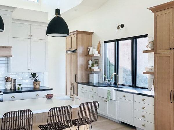 A modern kitchen with white cabinets, black countertops, a large island, and wicker bar stools, featuring pendant lights and a farmhouse sink under a large window.