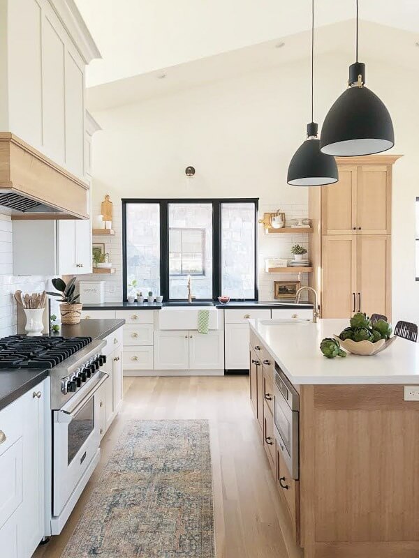 A modern kitchen with white cabinets, a large island, pendant lights, and a farmhouse sink under a window. The decor includes a patterned rug, open shelves, and a bowl of artichokes on the island.