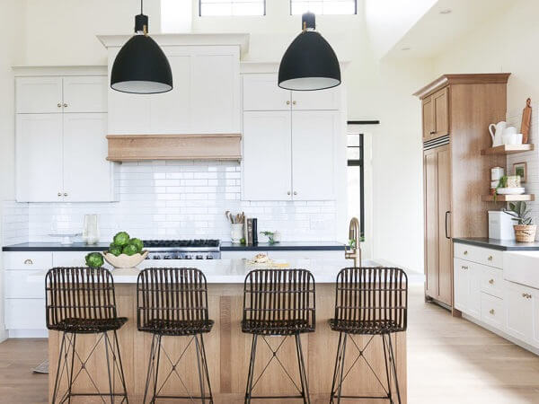 A modern kitchen with white cabinets, a large island with wicker bar stools, pendant lights, and a gas stove against a white subway tile backsplash.