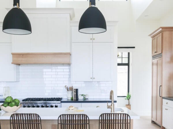 A modern kitchen with white cabinets, a large island with wicker bar stools, pendant lights, and a gas stove against a white subway tile backsplash.