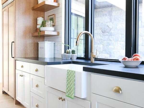 A modern kitchen corner featuring a farmhouse sink with a brass faucet, black countertops, white cabinets, and open shelves with decorative items.