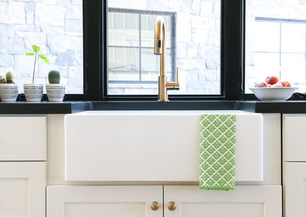 A close-up of a modern farmhouse sink with a brass faucet, set against a black countertop and a large window, with small potted plants and a bowl of fruit nearby.