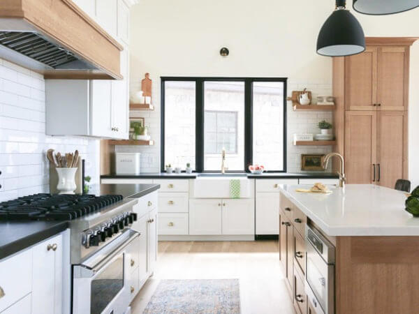 A modern kitchen with white cabinets, a black countertop, a large island, and a farmhouse sink under a window, featuring a gas stove and open shelves with decorative items.