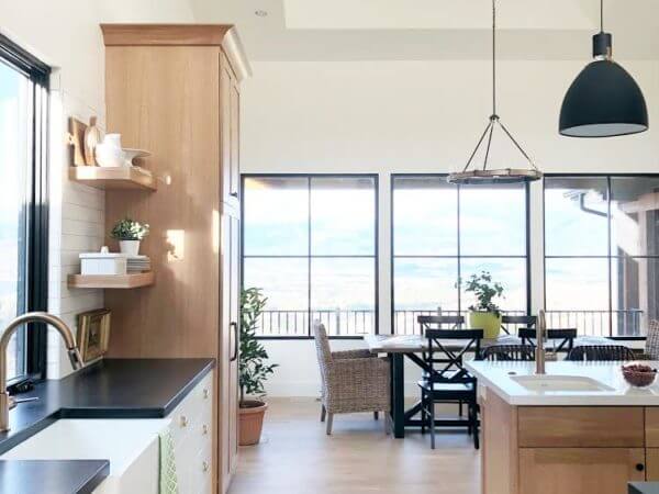 A modern kitchen with an island and pendant lights, opening into a dining area with a large wooden table, wicker chairs, and large windows providing natural light.