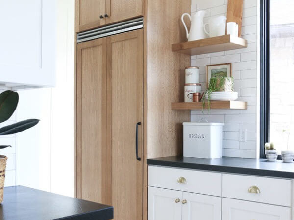 A modern kitchen corner with white cabinets, a black countertop, wooden pantry doors, and open shelves holding decorative items and a bread box.