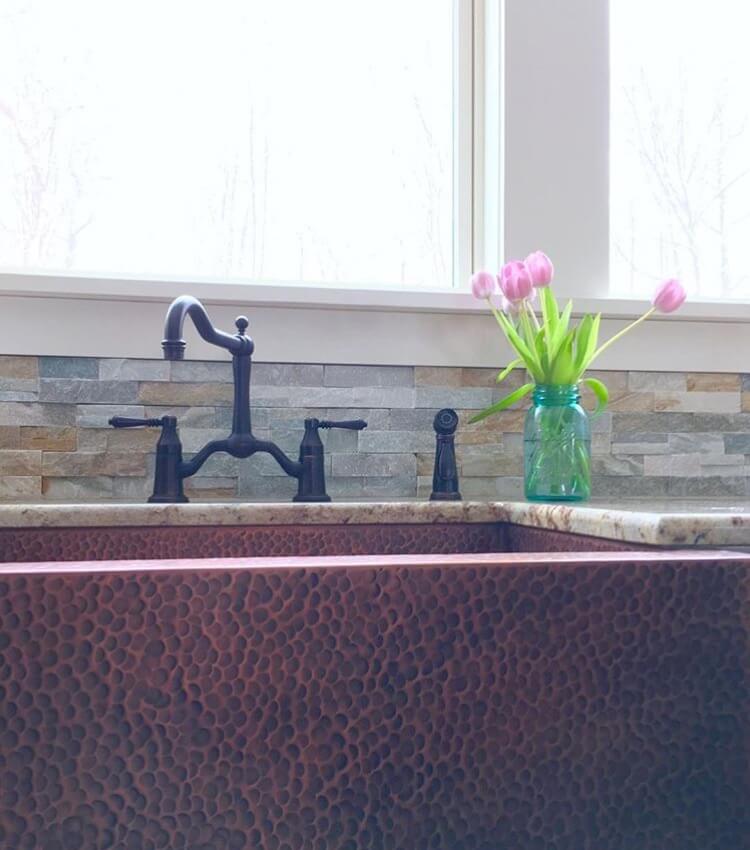 A close-up of a copper farmhouse sink with a textured finish, a black faucet, and a vase of pink tulips on the countertop, set against a stone tile backsplash.