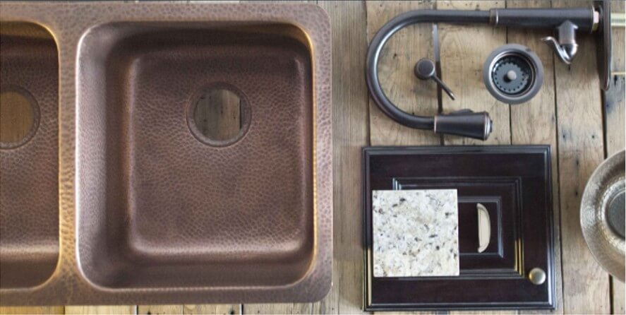 A top view of a copper double kitchen sink with a textured finish, accompanied by a black faucet, a sample granite countertop, and a dark wood cabinet door, all set on a wooden surface.