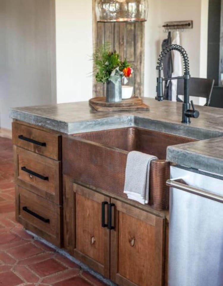 A rustic kitchen island with a copper farmhouse sink, a black spring faucet, concrete countertops, and wooden cabinetry.