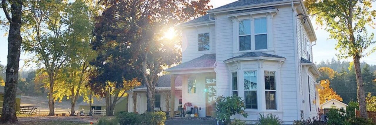 A beautiful white two-story house surrounded by tall trees, with sunlight filtering through the leaves, creating a warm and inviting atmosphere.