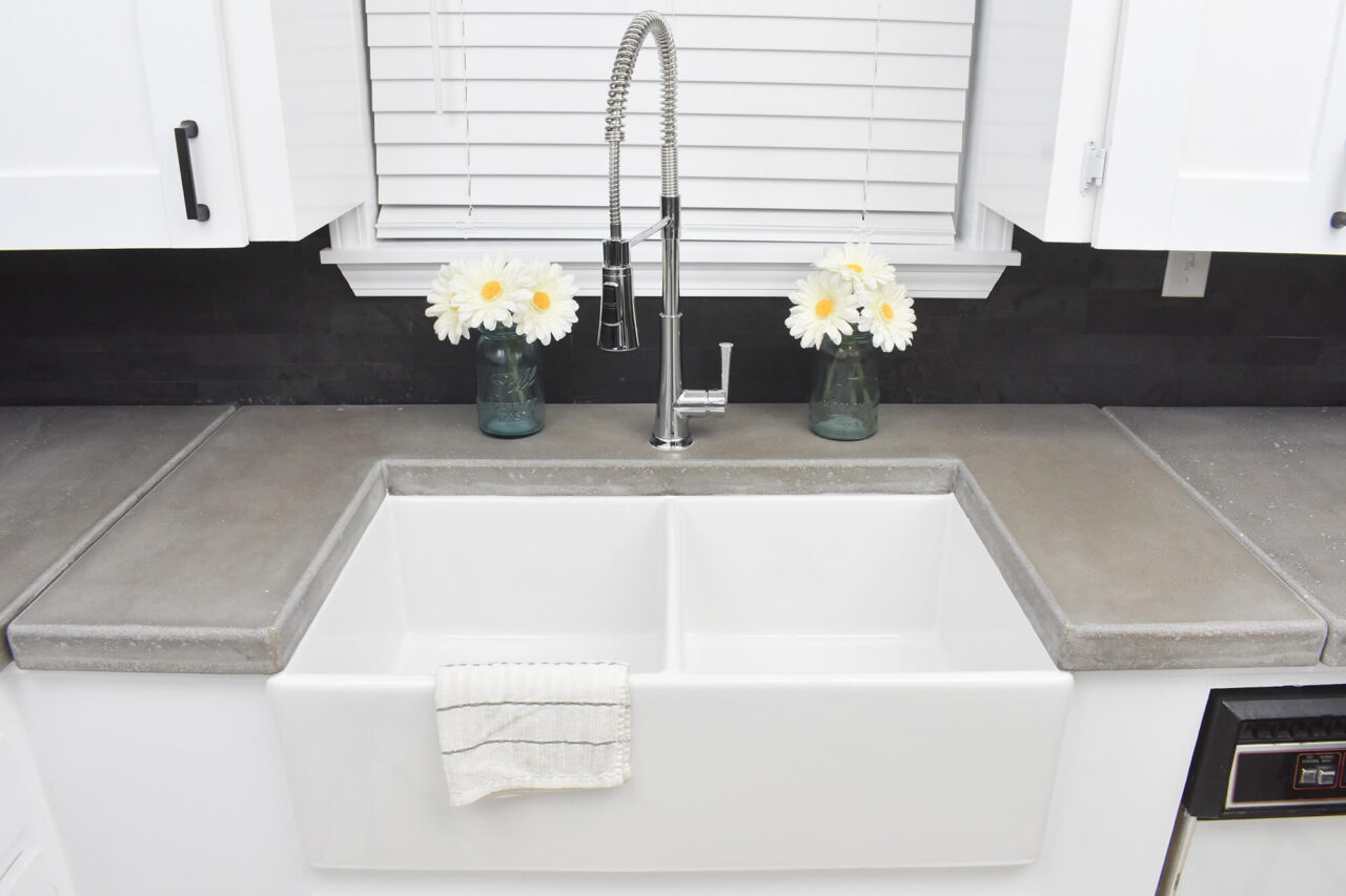 A white farmhouse sink with a spring faucet, set against concrete countertops and a dark backsplash, with two mason jars holding white daisies on either side.