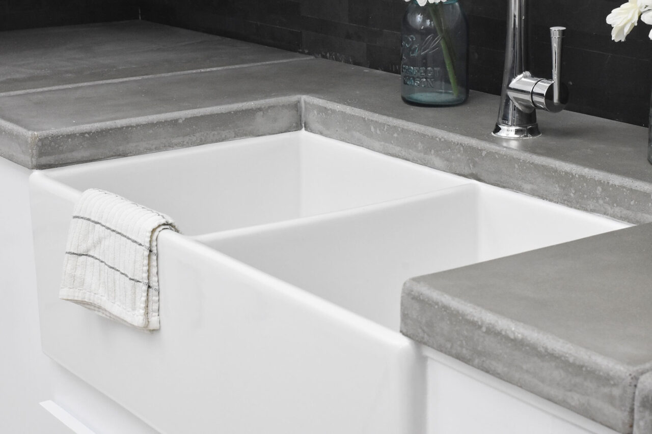A close-up of a white farmhouse sink with concrete countertops and a striped dish towel hanging over the sink's edge, set against a dark backsplash.