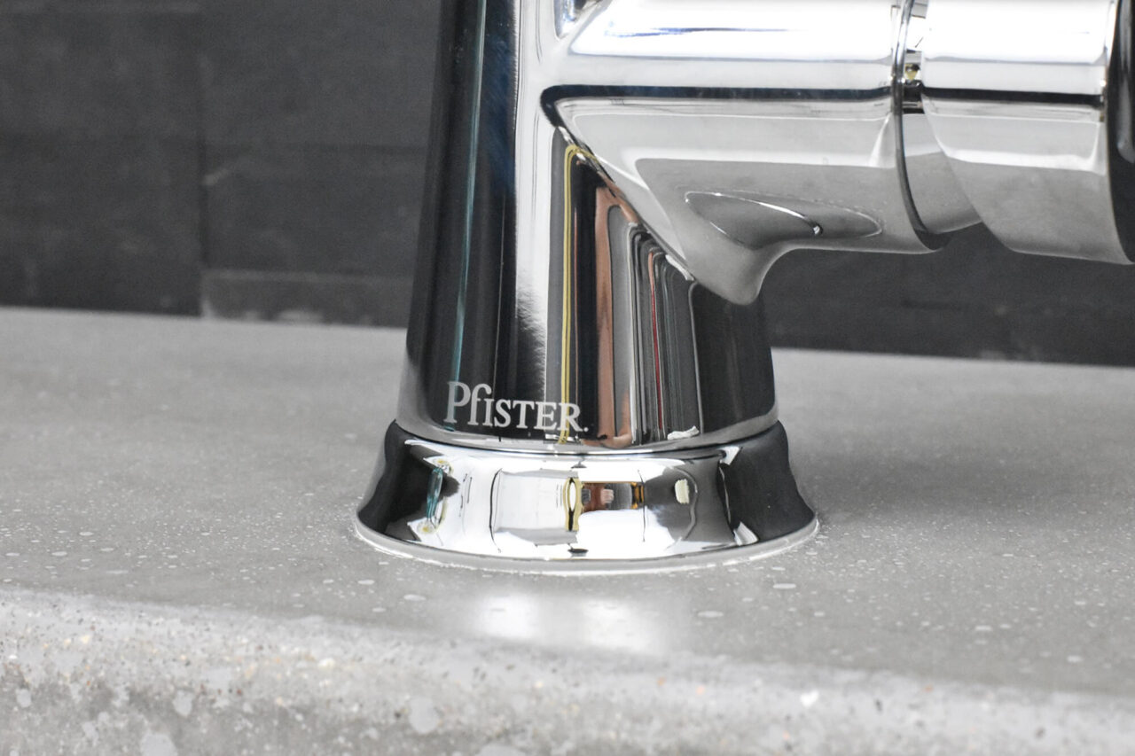 A close-up of the base of a chrome faucet with the brand name "Pfister" on a concrete countertop.