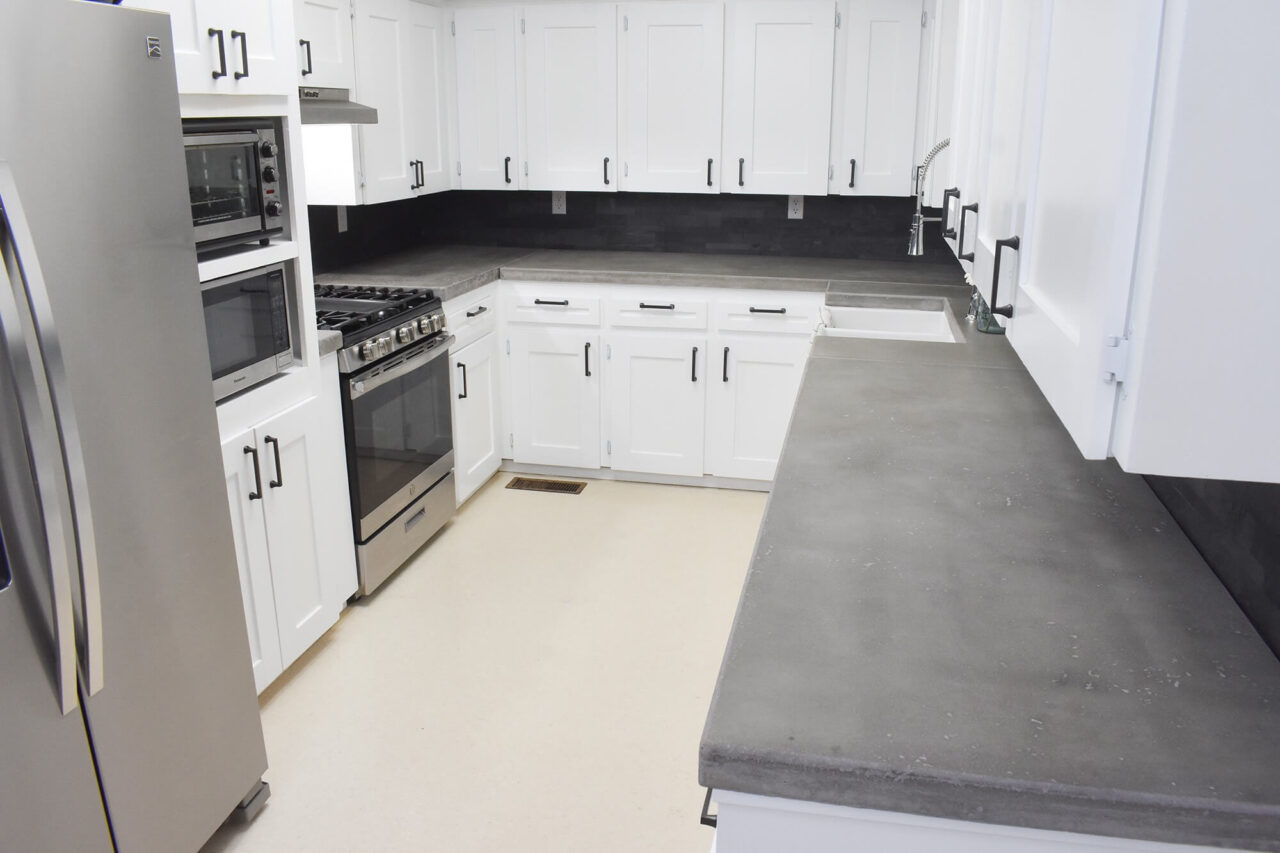 A modern kitchen with white cabinets, concrete countertops, stainless steel appliances, and a black backsplash.