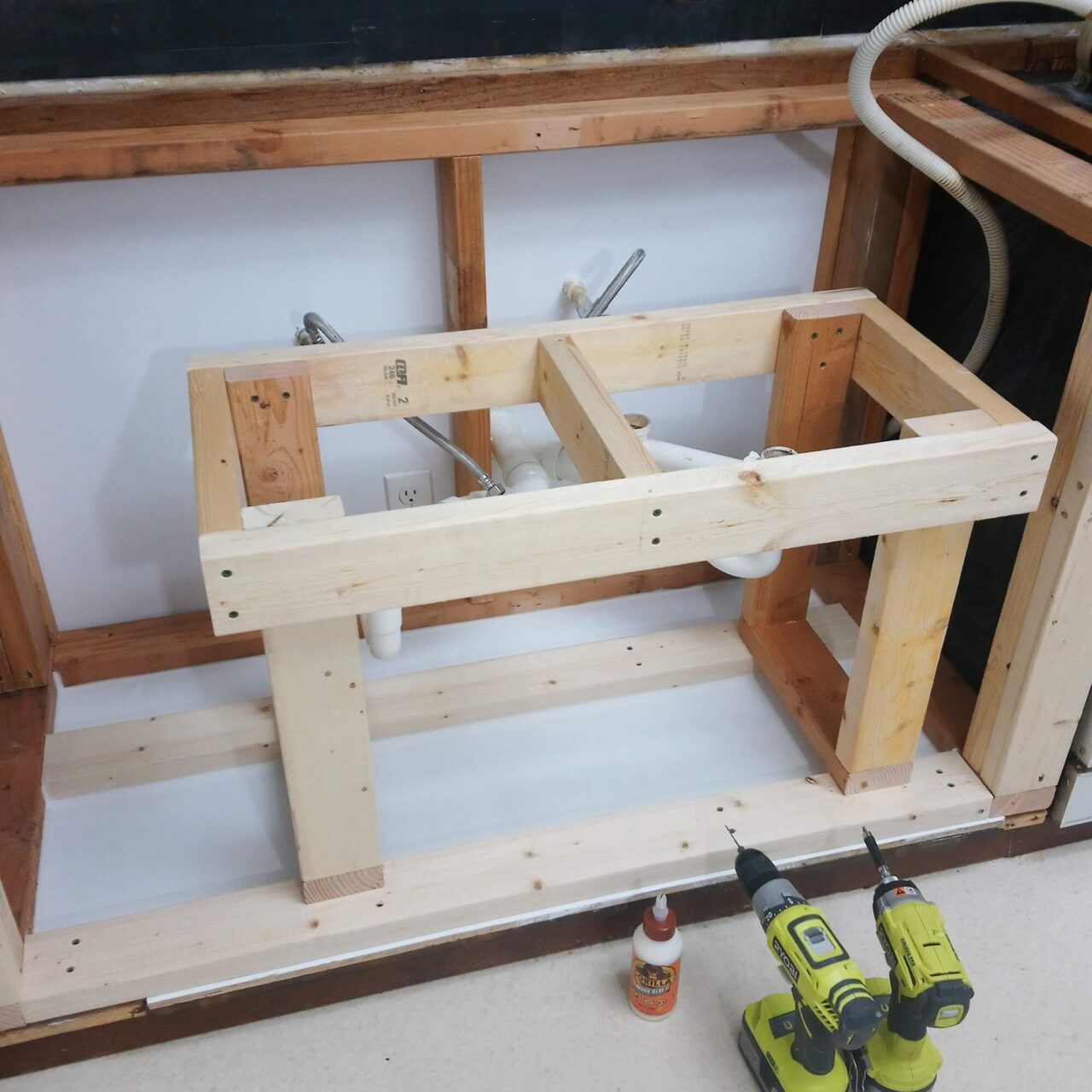 A wooden frame for a kitchen sink installation, with plumbing visible, set against an unfinished wall, alongside two yellow power drills and a bottle of wood glue.