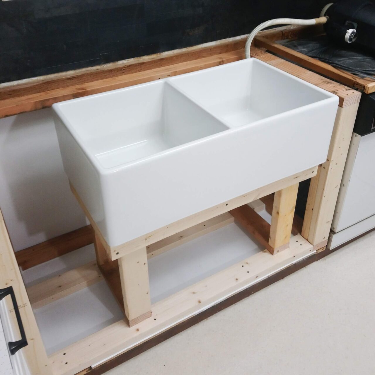 A white double farmhouse sink being installed into a wooden frame, with plumbing visible in an unfinished kitchen area.