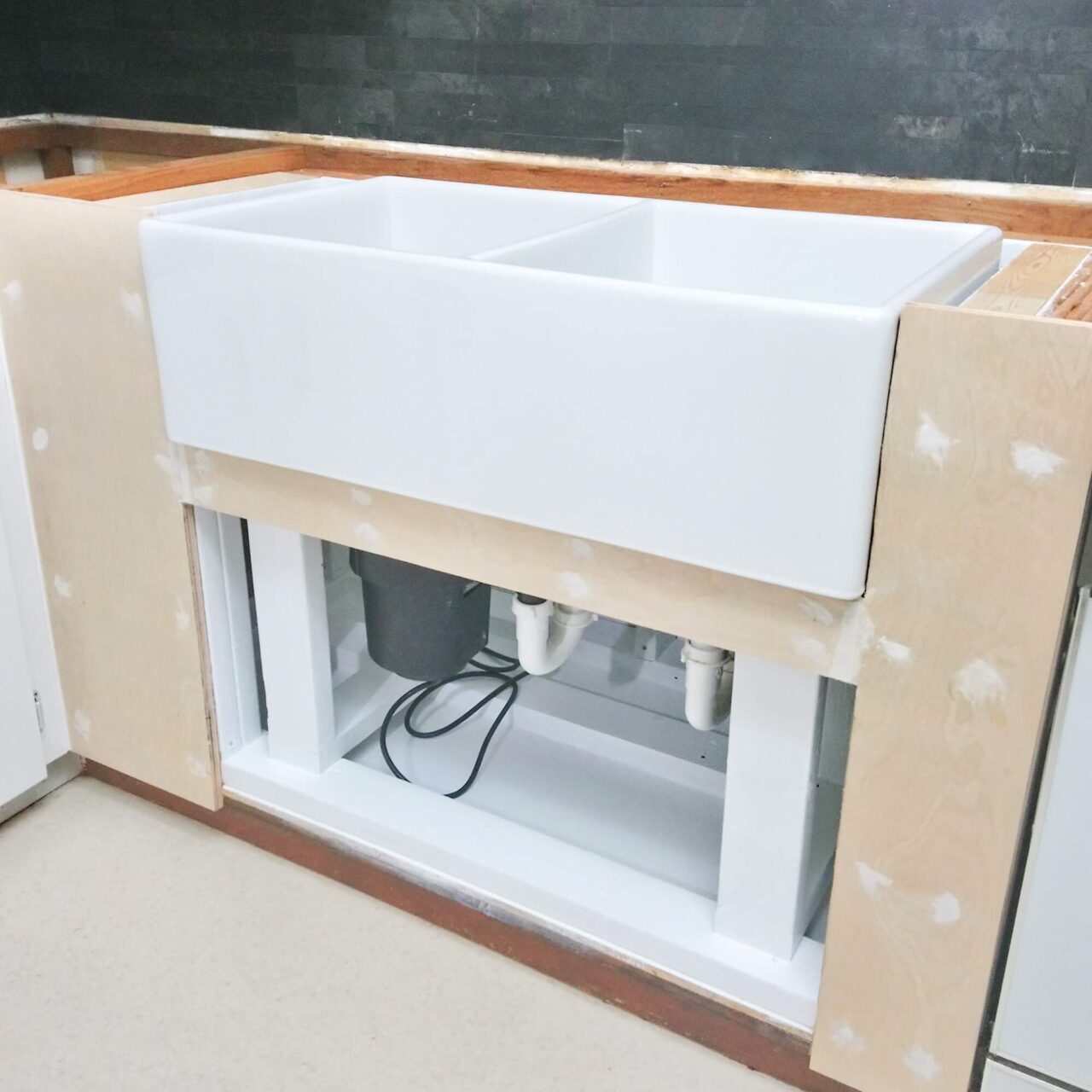 A white double farmhouse sink partially installed into a wooden frame, with visible plumbing and a garbage disposal underneath, in an unfinished kitchen area.