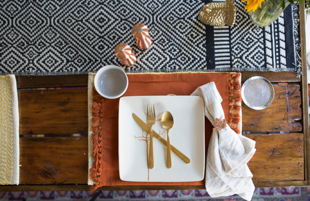 A place setting on a wooden table with a patterned table runner, featuring a white square plate, gold utensils, and a white napkin with a decorative ring.