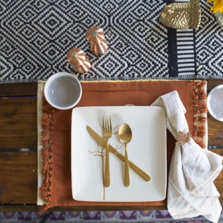 A place setting with a white square plate, gold utensils, a white napkin with a decorative ring, and a small bowl on a patterned table runner.