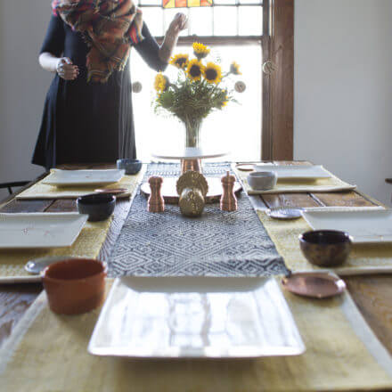A dining table set with plates, bowls, and a centerpiece of sunflowers, with a person standing near the window.
