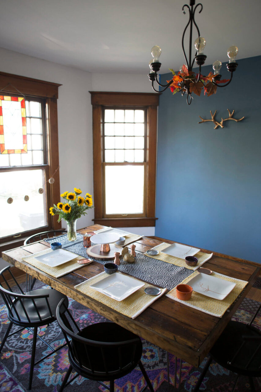 A dining room with a wooden table set for six, featuring a sunflower centerpiece, decorative plates, and a patterned table runner.