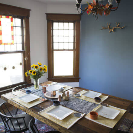 A dining room with a wooden table set for six, featuring a sunflower centerpiece, decorative plates, and a patterned table runner.