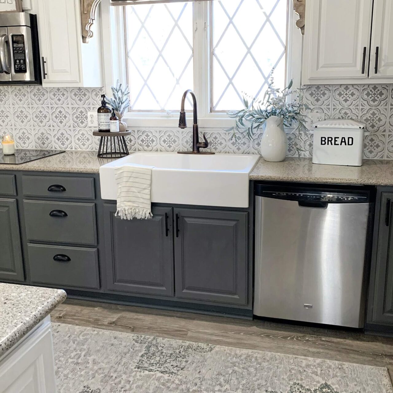 A stylish kitchen with a white farmhouse sink, dark gray cabinets, a patterned tile backsplash, and a stainless steel dishwasher, featuring decorative items like a "BREAD" box and a vase of greenery.