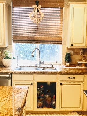 Cozy kitchen with cream-colored cabinets, a double sink under a large window with bamboo blinds, granite countertops, and a decorative chandelier above.