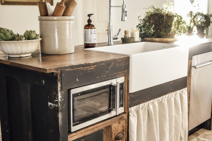 A completed kitchen island with a white farmhouse sink, microwave, and rustic wooden cabinetry, decorated with kitchen items and plants.