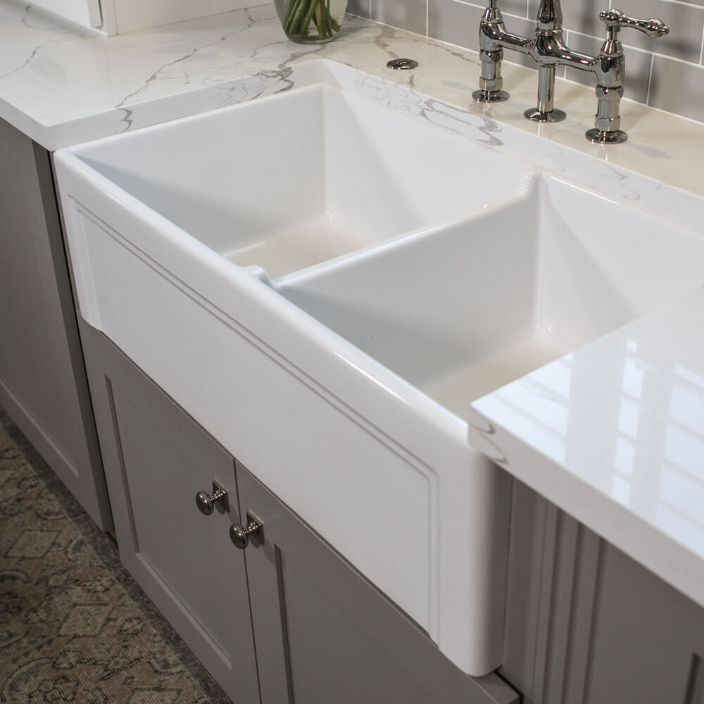 A close-up of a white double-basin farmhouse sink with a marble countertop and chrome faucet, set against gray cabinetry.