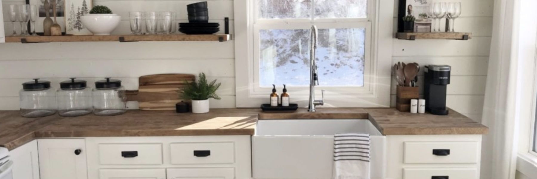 A bright kitchen with white cabinets, a farmhouse sink, and wooden countertops. Shelves above and around the sink hold dishes, decor, and a coffee maker. Large windows provide natural light and a view outside.