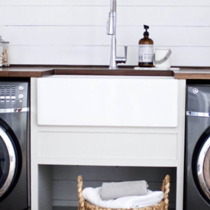 Laundry room setup with a white farmhouse sink, wooden countertop, stainless steel faucet, and a wicker basket filled with towels, flanked by a washer and dryer.