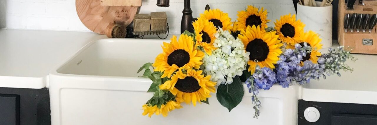 Farmhouse sink with a bouquet of sunflowers, white hydrangeas, and purple flowers, alongside kitchen utensils and a bronze faucet.