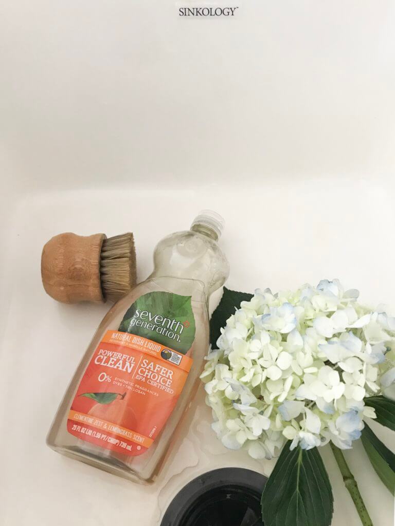 Bottle of Seventh Generation dish soap, a scrub brush, and white hydrangea in a farmhouse sink.