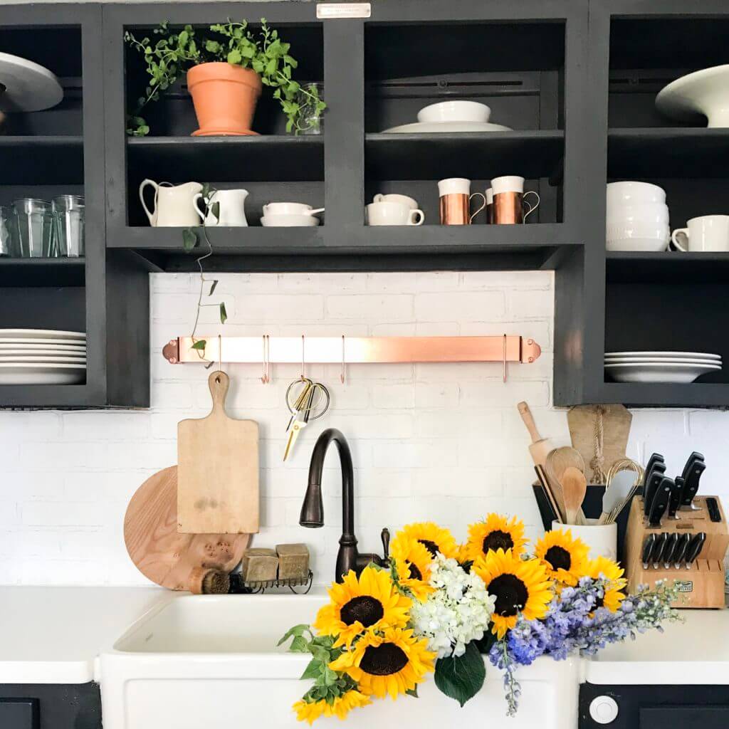 Farmhouse kitchen with black open cabinets, bronze faucet, and a sink adorned with sunflowers, white hydrangeas, and purple flowers.