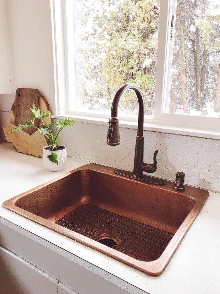 A kitchen sink area with a hammered copper sink, a dark faucet, a large window with a snowy view, and a small plant on the white countertop. A wooden cutting board is in the corner.