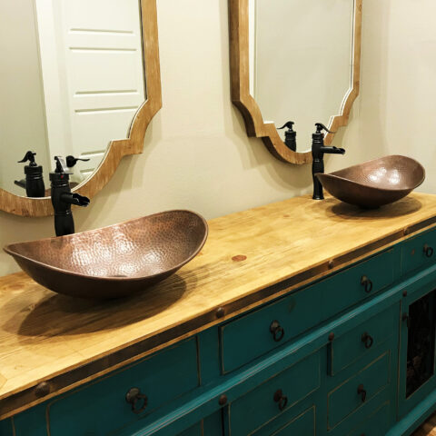 A duo of hand hammered copper vessel bathroom sinks in a teal cabinet and light wooden top in a bathroom.
